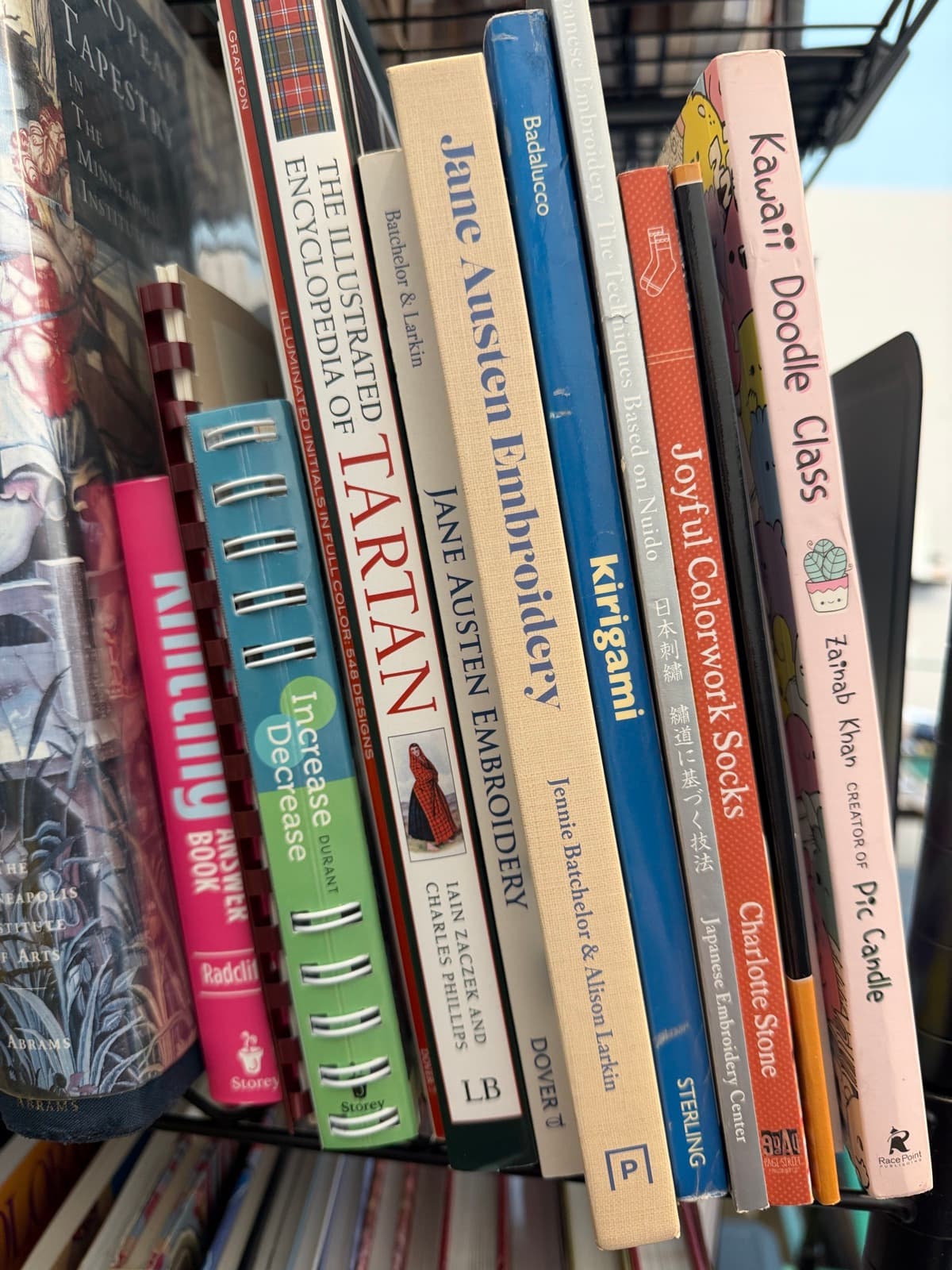 Book spines in the Fiber Arts Houston library, including Japanese embroidery, tartan, colorwork sock, and Jane Austen embroidery titles
